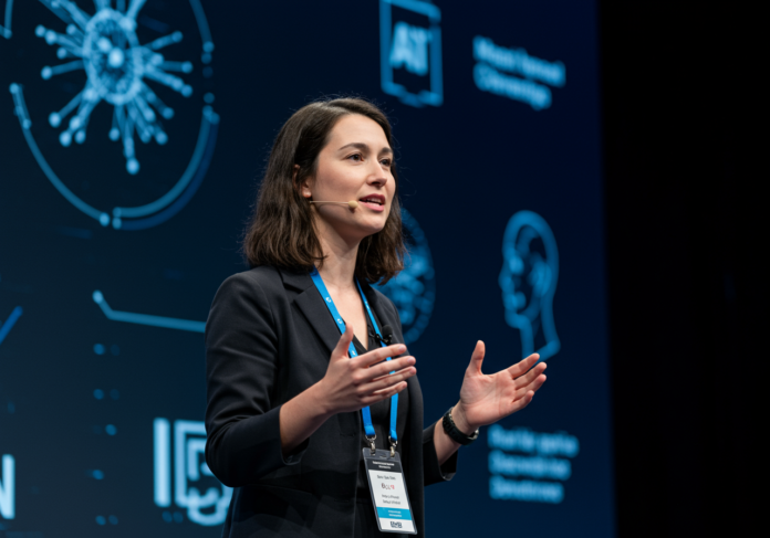 Uma carismática cientista cognitiva palestrando com paixão em um palco bem iluminado numa importante conferência de Inteligência Artificial. Ao fundo, são sutilmente apresentadas visualizações abstratas de redes neurais e elementos de interação humano-computador. A palestrante é o foco central da imagem, transmitindo especialização e inovação.